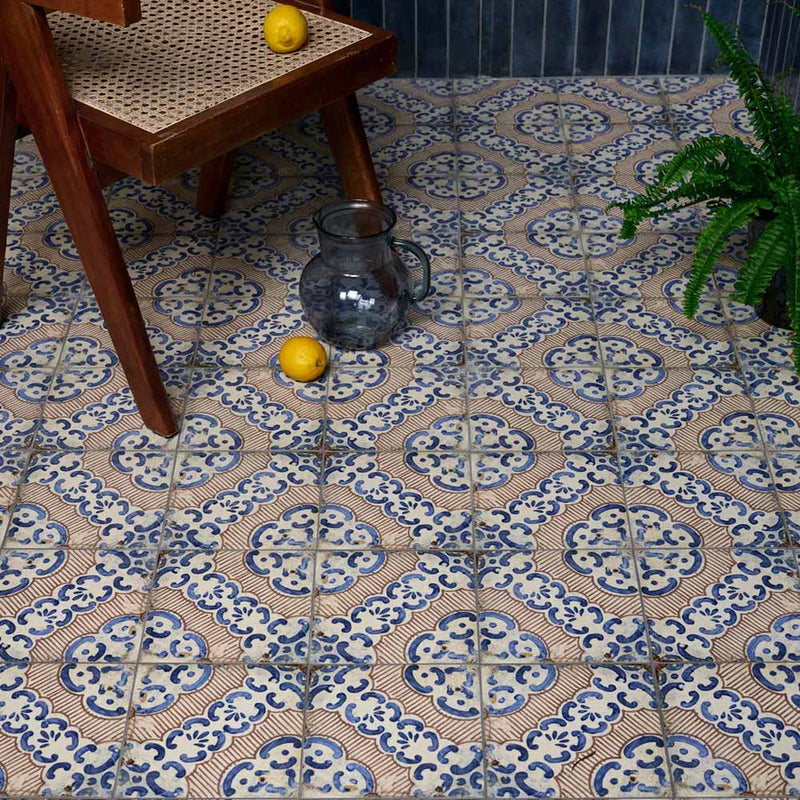 Image of Sardinia Masseria Pattern tile 20x20cm on a kitchen floor. Wooden chair to the left with a blue glass jug on the floor next to a lemon, blue gloss wall tiles and a green plant on the floor.