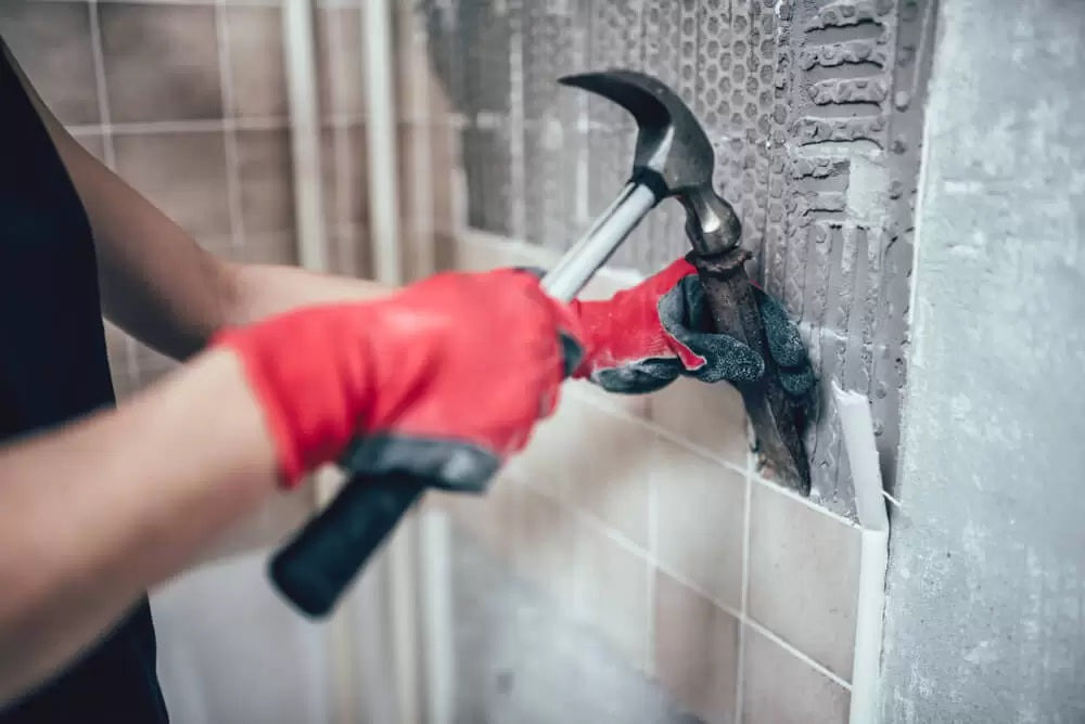 person with safety gloves, hammer and chisel removing tiles from wall 
