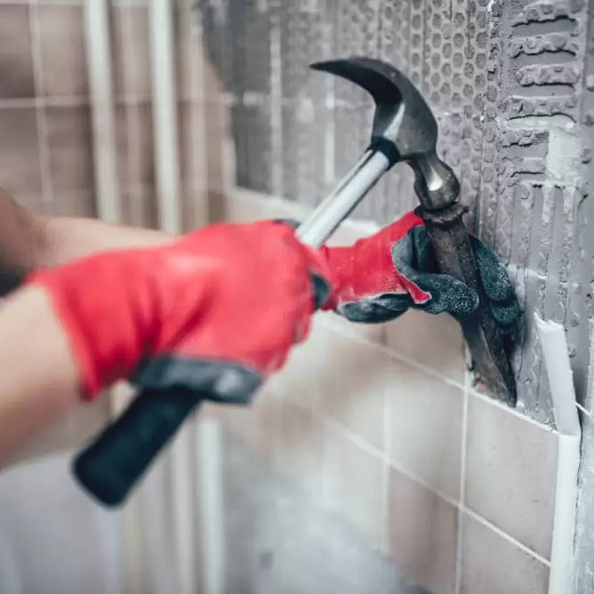 person with safety gloves, hammer and chisel removing tiles from wall 