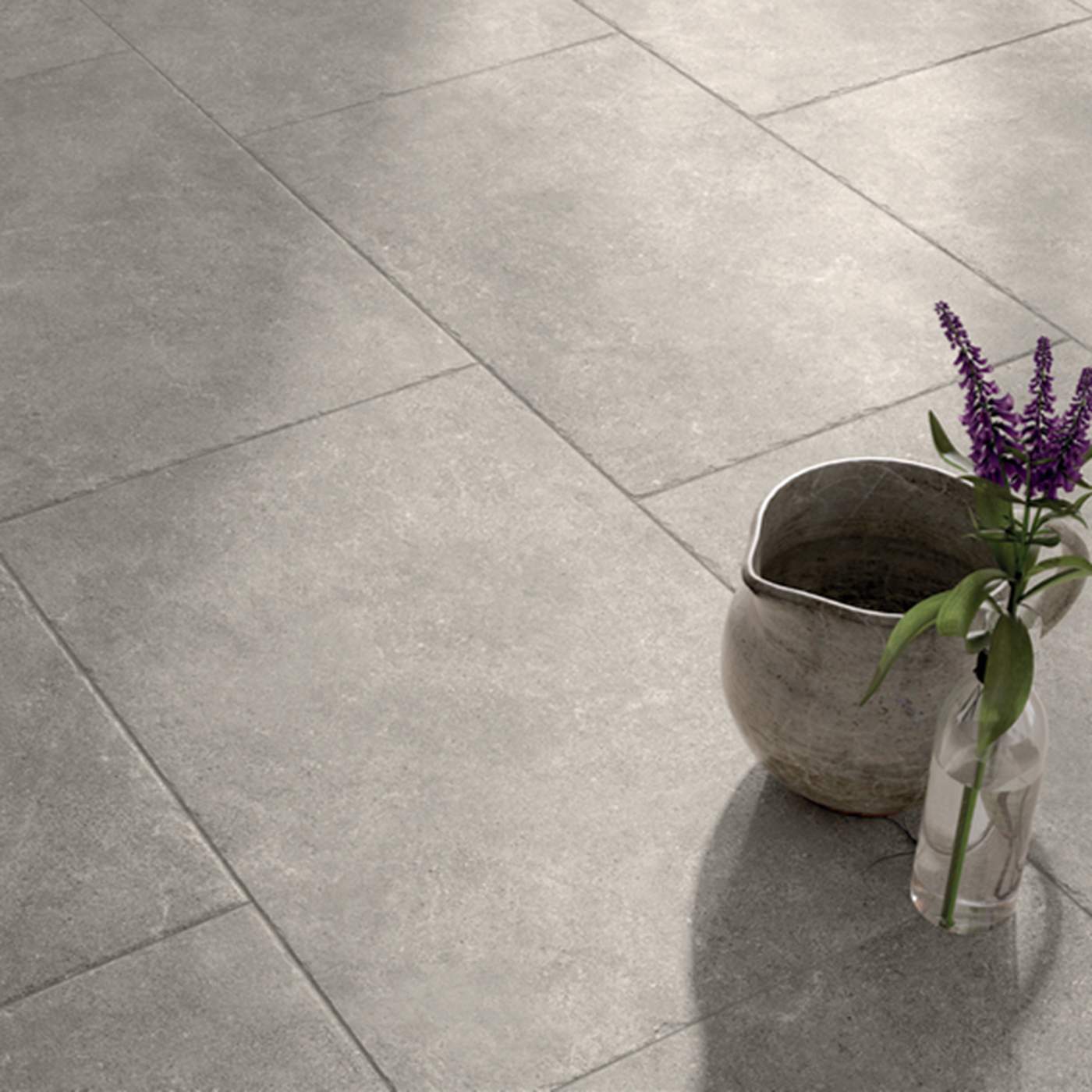 Image of Large square grey stone-look floor tiles with a matte textured finish, styled with a rustic ceramic pitcher and a small glass vase containing purple flowers in the lower right corner.