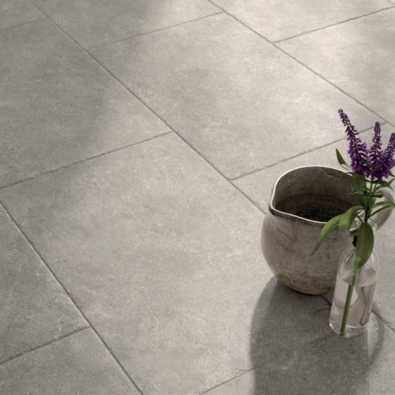 Image of Large square grey stone-look floor tiles with a matte textured finish, styled with a rustic ceramic pitcher and a small glass vase containing purple flowers in the lower right corner.
