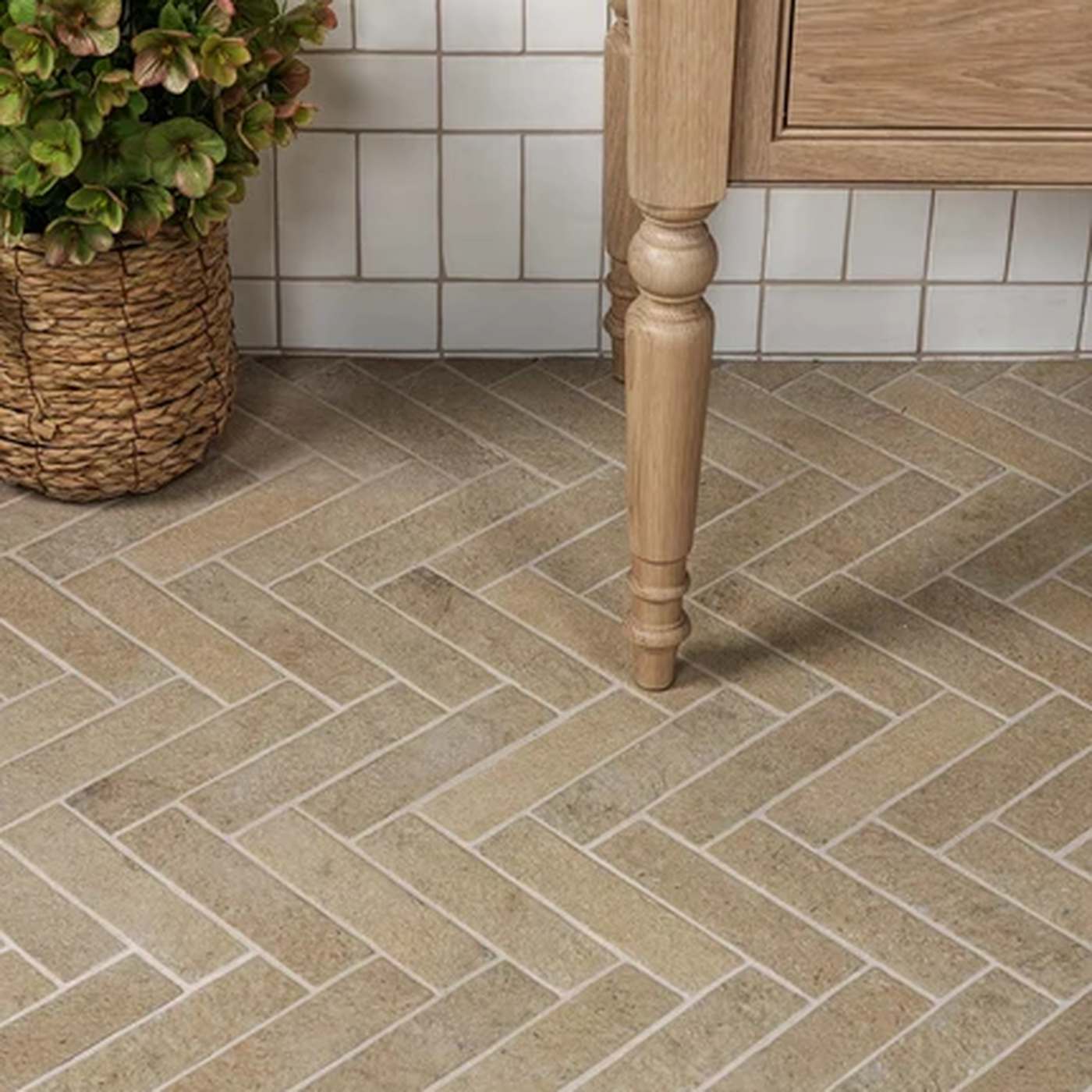 Image of Beige herringbone stone-look floor tiles installed in a kitchen setting, shown with a wooden cabinet leg and a potted plant.