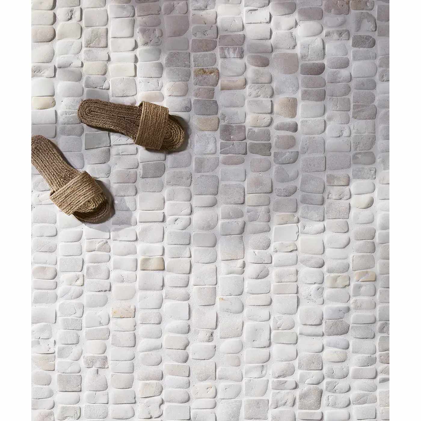 Image of A pair of natural jute slide sandals placed on a light gray and white pebble mosaic tile floor.