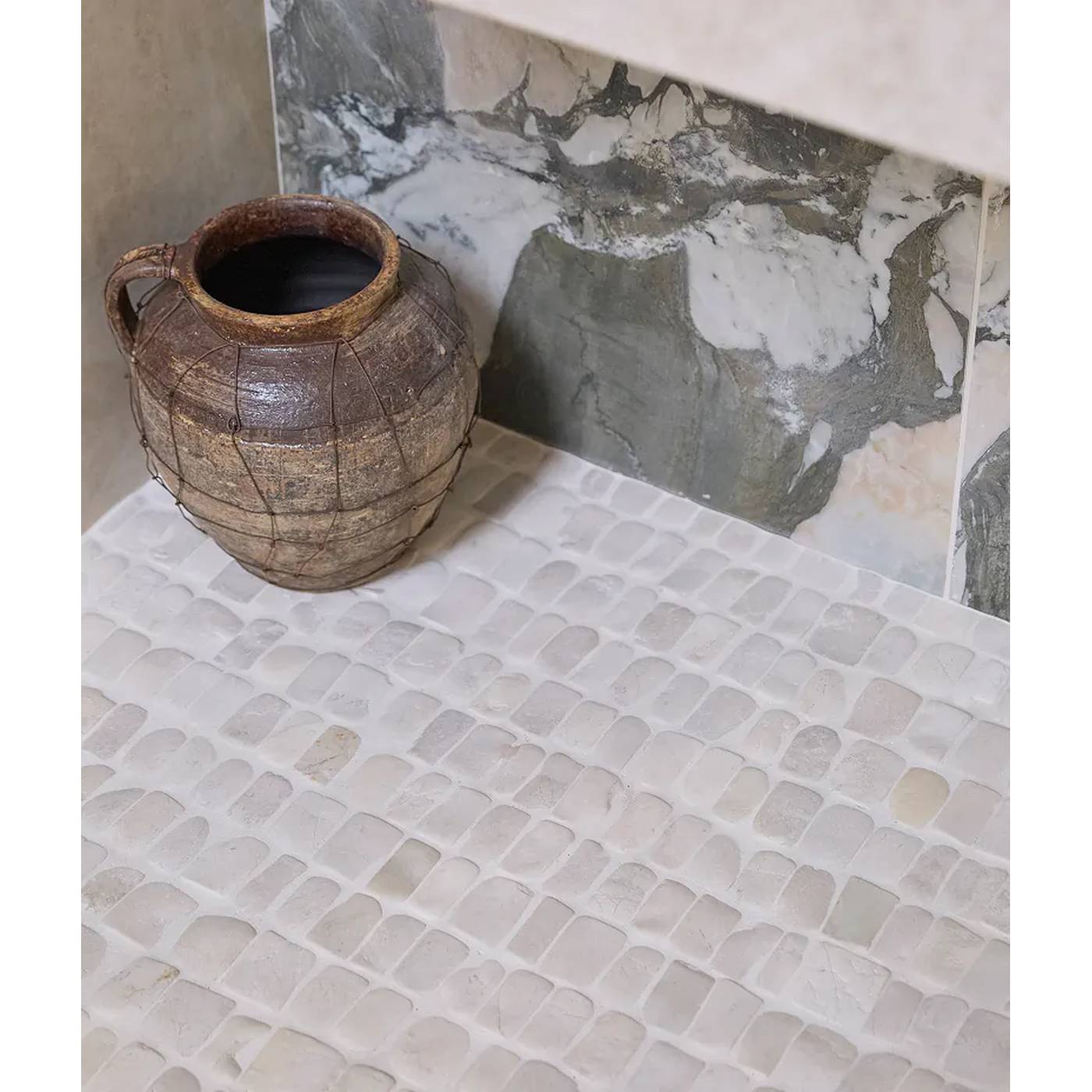 Image of A rustic brown ceramic pot with a wire handle, placed on a light beige pebble mosaic tile floor against a gray and white marble-look tile backsplash.