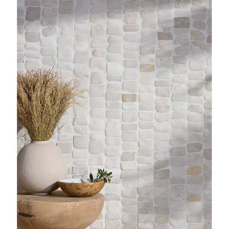 Image of A wall covered in rectangular stone mosaic tiles in warm off-white, beige, and light gray tones, styled with a round beige vase holding dried grass, a wooden bowl, and a small green plant on a wooden surface.