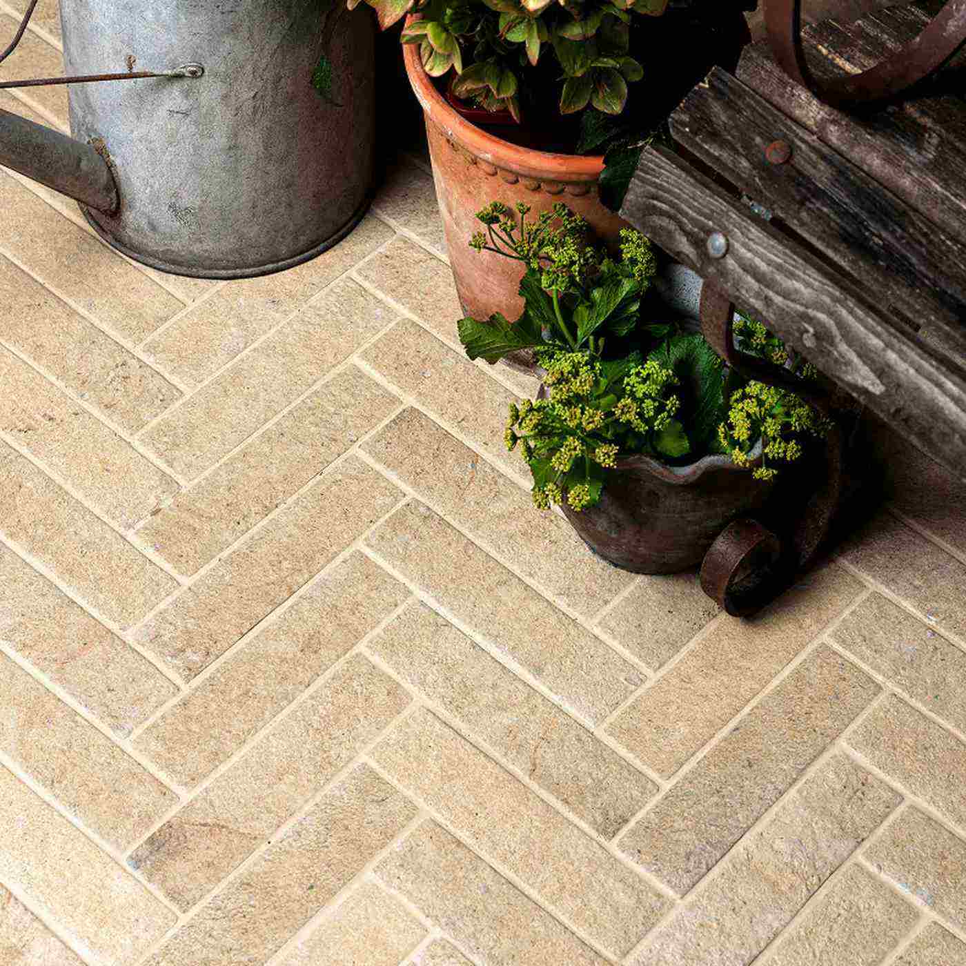 Image of Beige rectangular stone tiles arranged in a herringbone pattern, styled with potted plants and rustic garden props.