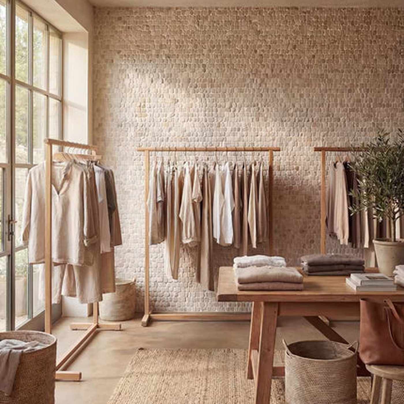 Image of A styled interior retail space with multiple wooden clothing racks holding neutral-toned garments, a wooden table with folded textiles, woven baskets, and a textured stone wall. No person or animal is present, but this is not a clean isolated product shot; it is a real environment, so subject_type is Lifestyle.
