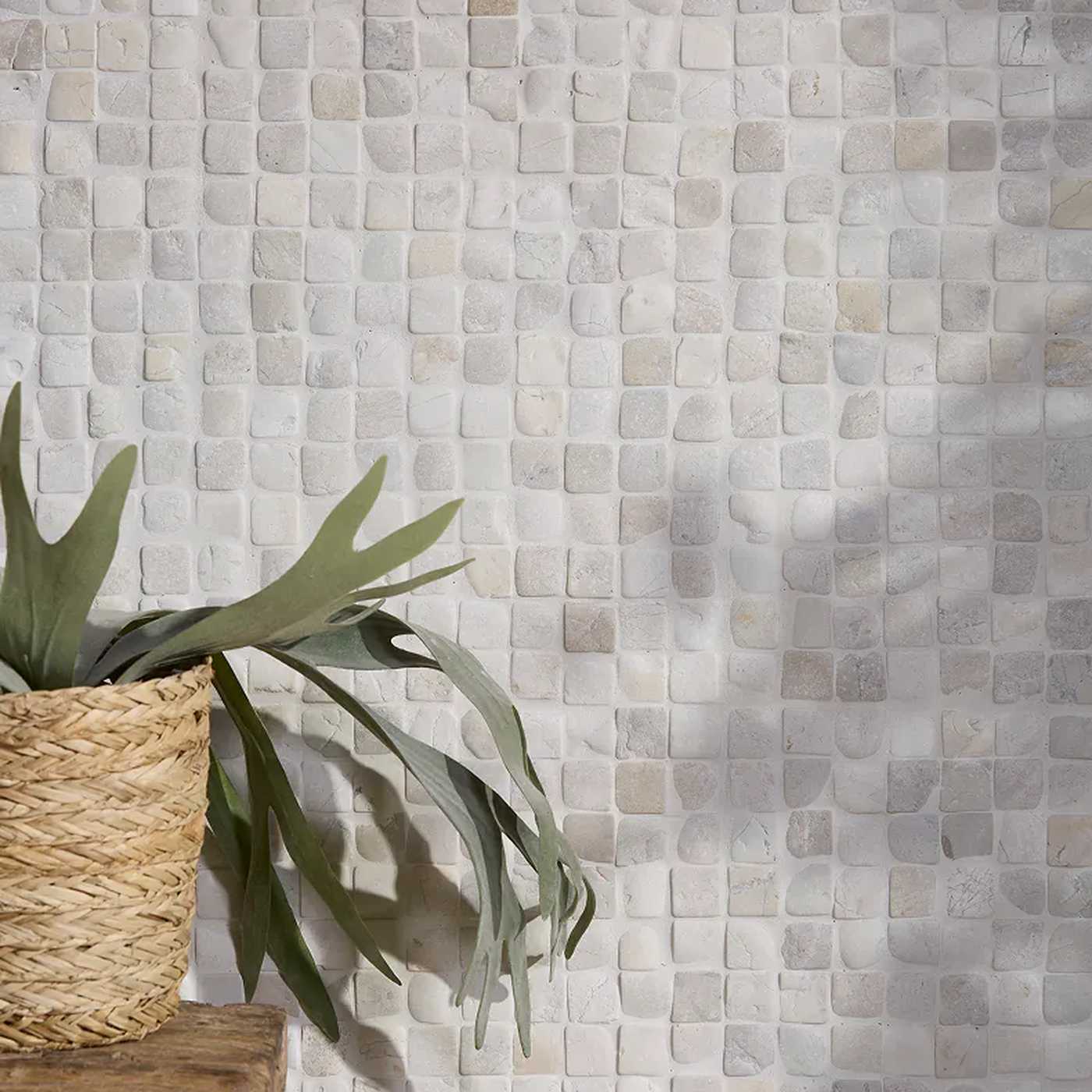 Image of Square mosaic wall tiles in a light beige and gray stone pattern, styled with a woven basket planter holding green leaves on a wooden ledge in the lower left.