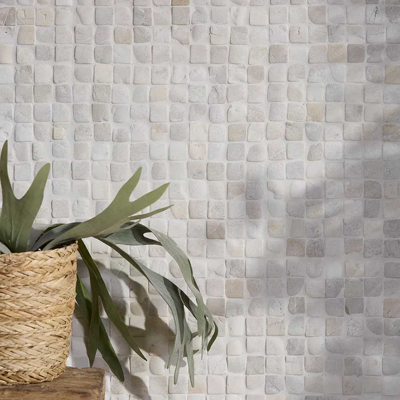 Image of Square mosaic wall tiles in a light beige and gray stone pattern, styled with a woven basket planter holding green leaves on a wooden ledge in the lower left.