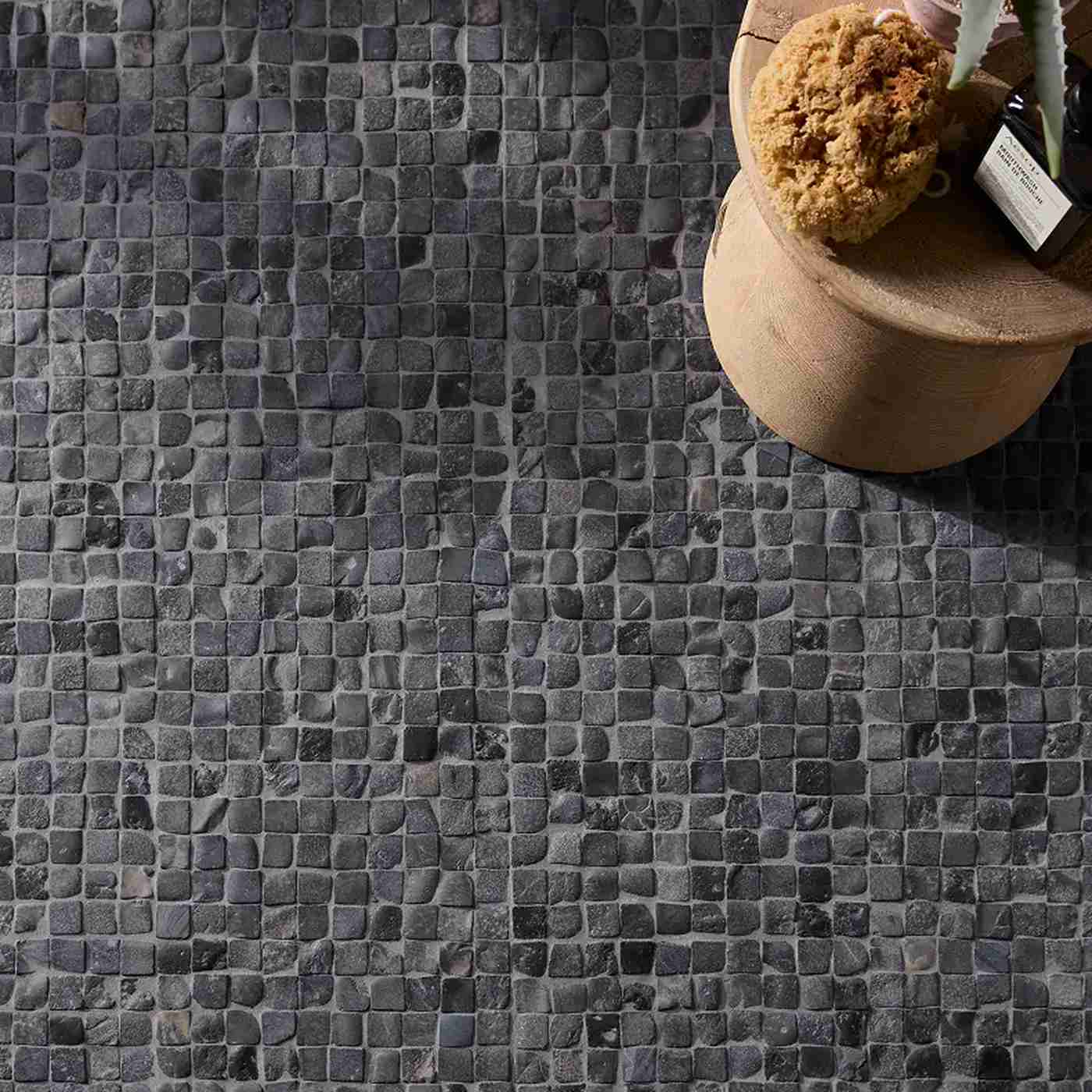 Image of Dark charcoal and black mosaic tile floor with a round wooden stool holding a sponge and a small bottle in the upper right corner.