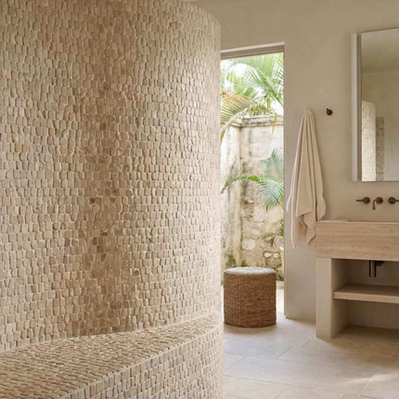 Image of A modern bathroom interior with a large cylindrical wall-mounted vessel or planter made of textured woven natural fibers, a matching built-in shelf, a stone vanity, a towel, mirror, and an open doorway revealing tropical greenery outside.