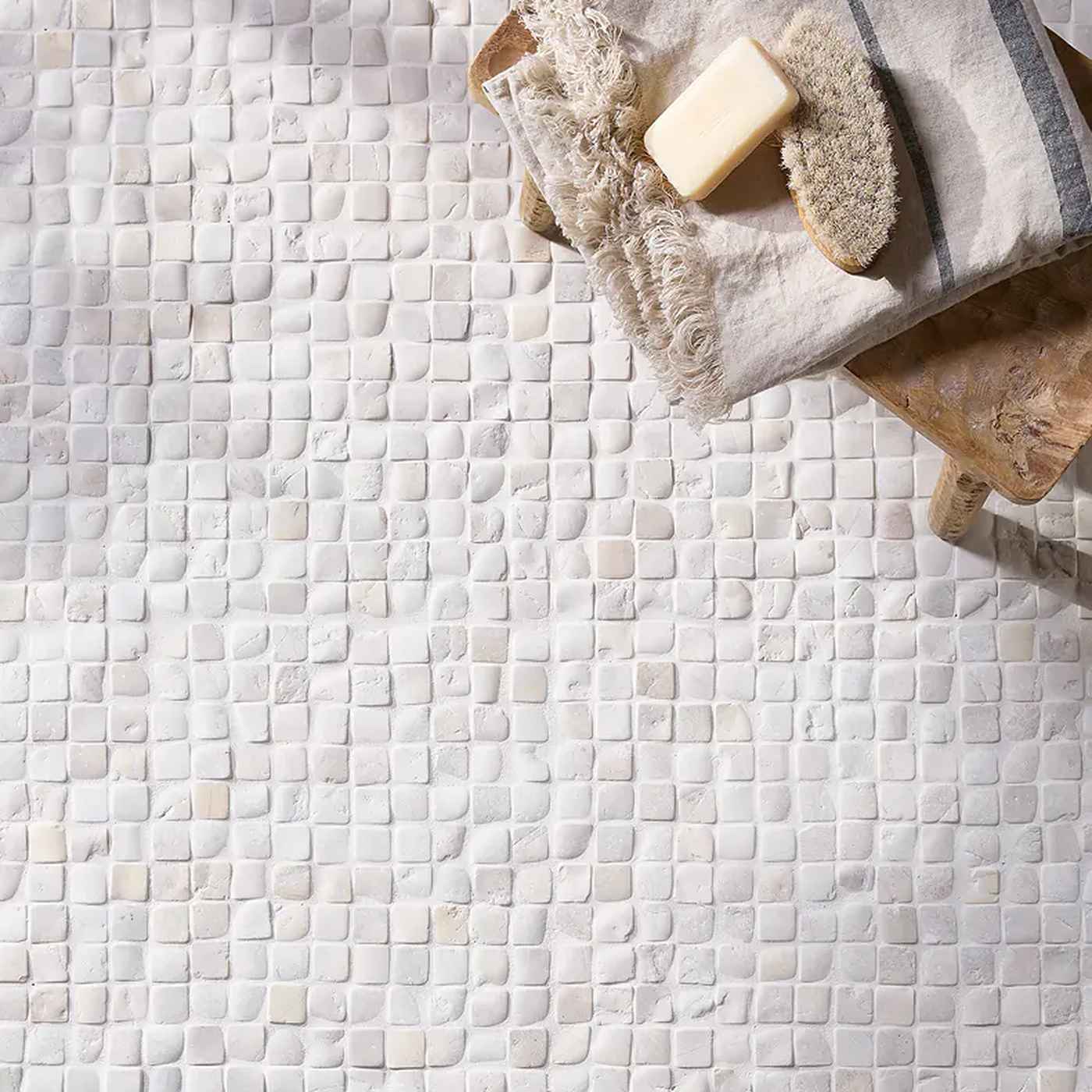 Image of White and beige mosaic tile floor with a folded cream towel, a bar of soap, and a natural bristle brush resting on a wooden stool in the upper right corner.