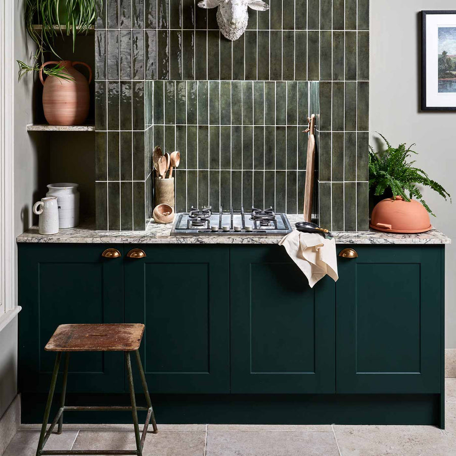 Foundry Smeraldo tile 6x24.5cm on a kitchen wall with dark green kitchen cabinets, marble worktop with terracotta potted plants and white jugs. Wooden, industrial stool sits on a stone tiled floor.