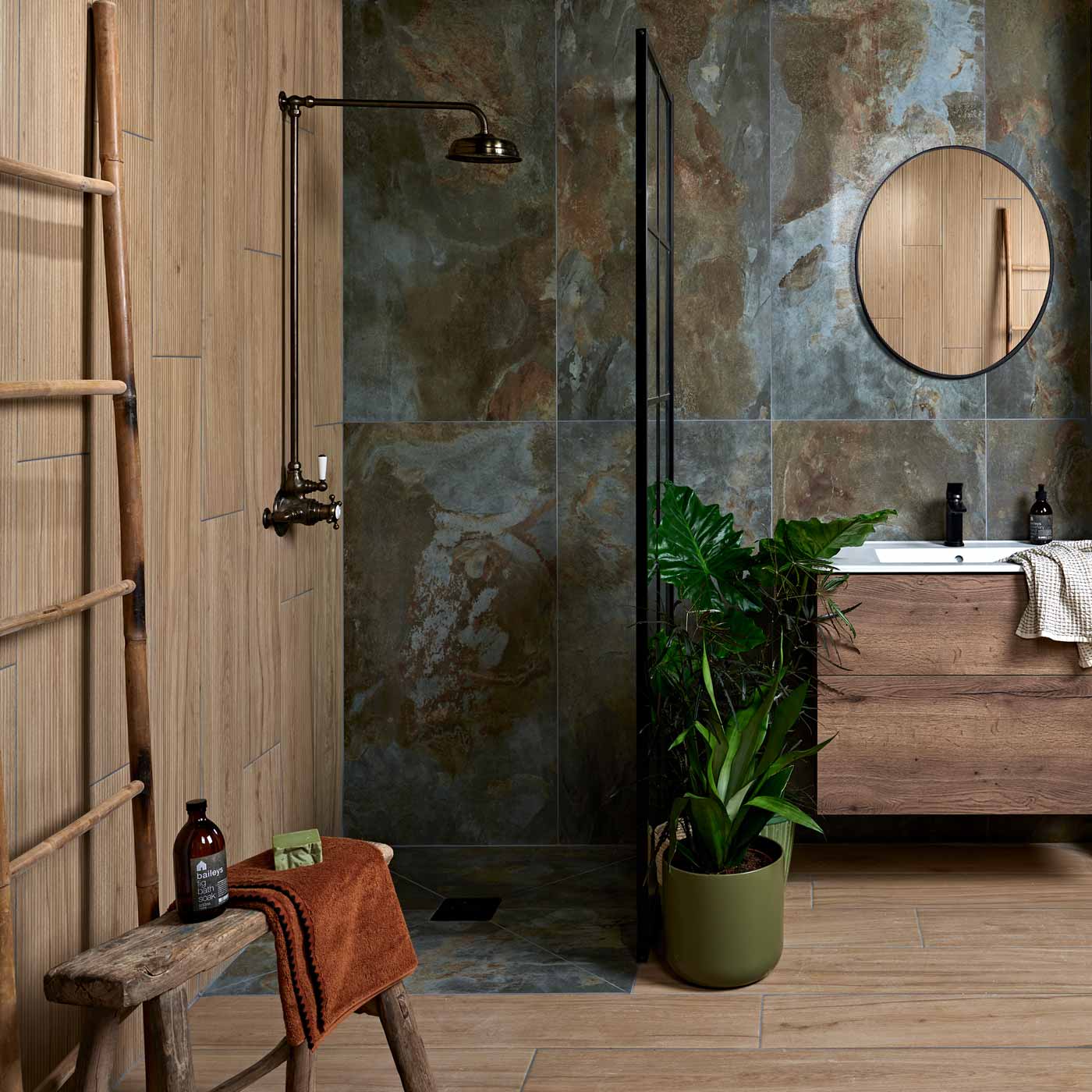 Komodo Matt Green tile 60x120cm in a bathroom setting. Wood effect tiles in a black metal shower, wooden vanity unit with white rectangular sink. Oval shaped mirror above that. Wood effect tiles on the floor, with green potted floor plants, wooden long stool with toiletries and towel on.