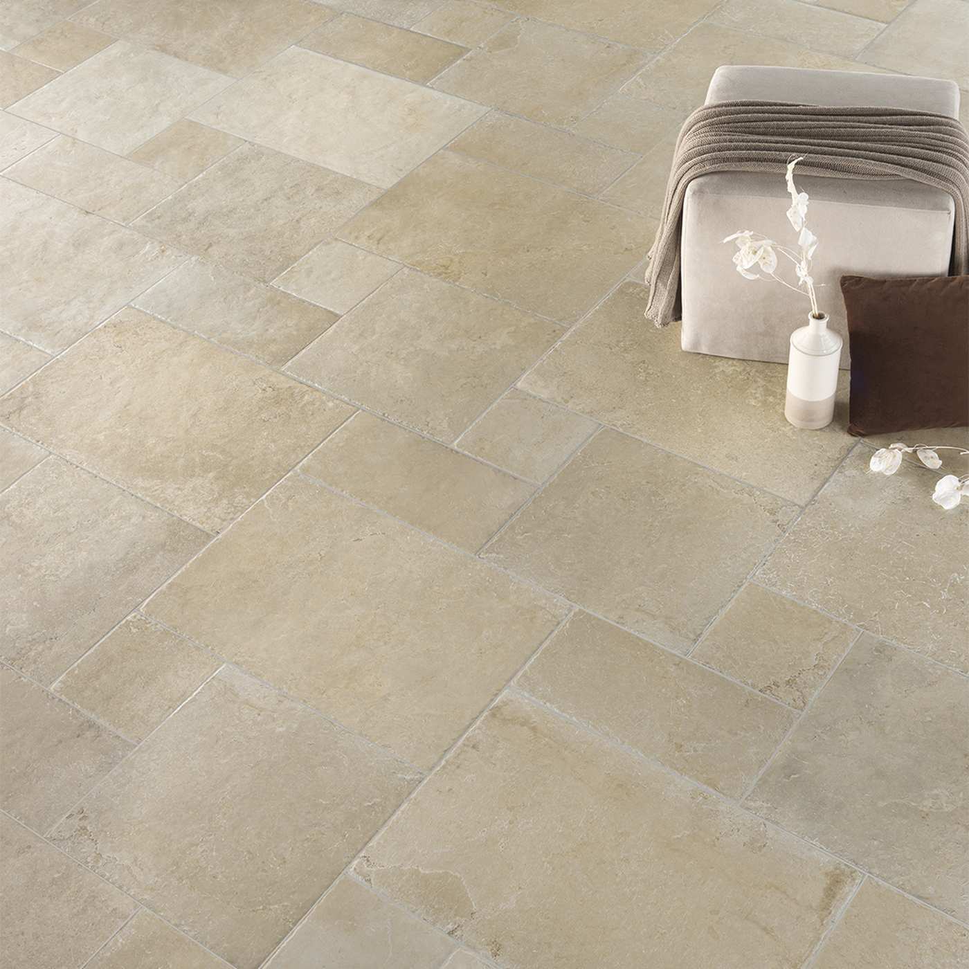Image of Beige stone-look rectangular floor tiles arranged in a herringbone pattern, shown in a styled interior scene with a small upholstered bench, draped throw, vase, and cushions.