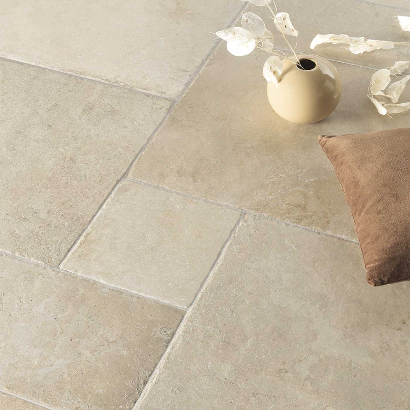 Image of Beige rectangular stone-look floor tiles arranged in a herringbone pattern, styled with a small cream vase holding dried white flowers and a brown cushion in the corner.