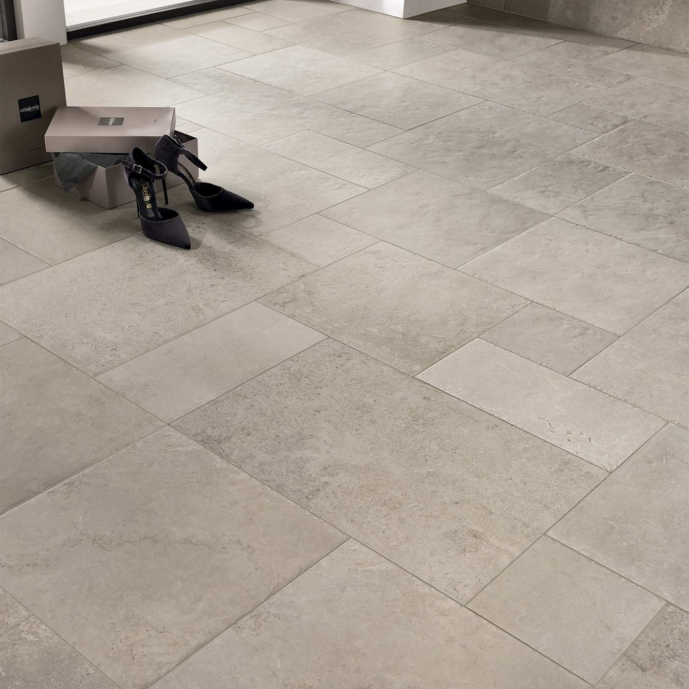 Image of Large light grey stone-look floor tiles arranged in a herringbone pattern, with a small bench, shoe box, and black high heels placed in the upper left corner.