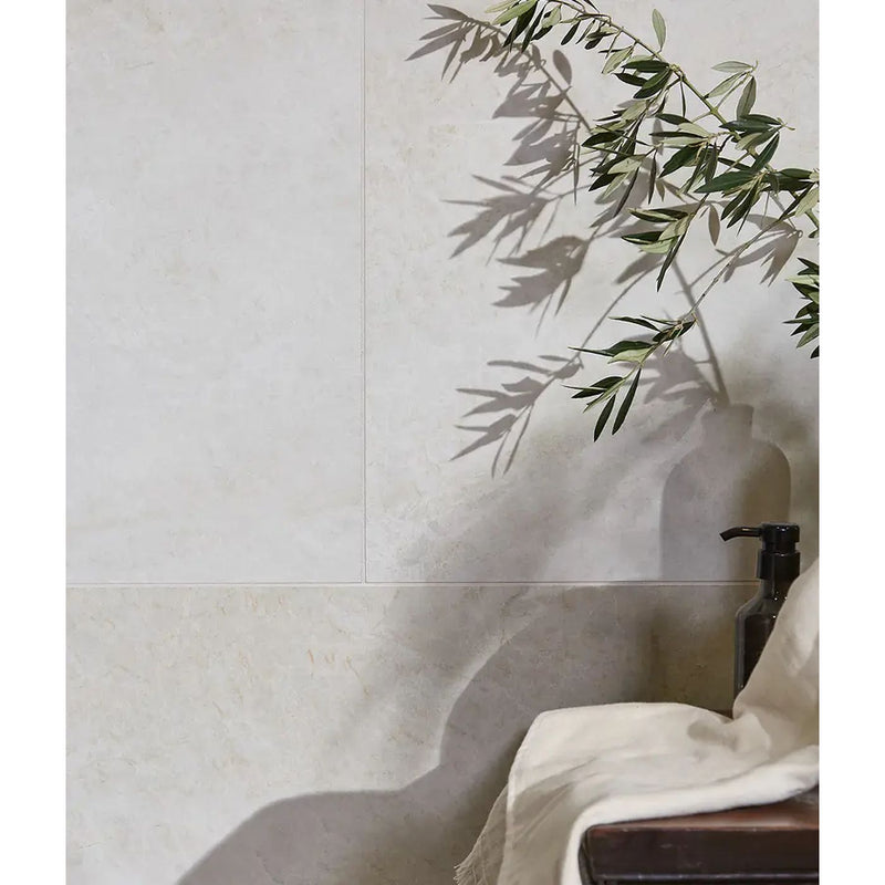 Image of Beige textured wall tiles with a large shadow cast by olive branch leaves, next to a dark glass bottle and draped white fabric.