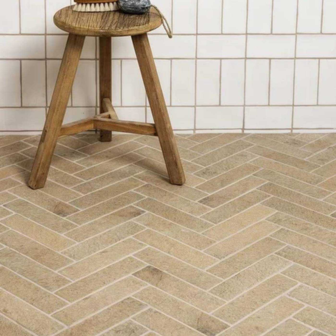 Image of Beige herringbone-patterned porcelain or ceramic floor tiles with white grout lines, shown in a bathroom setting with a wooden stool and white subway tiles on the wall.