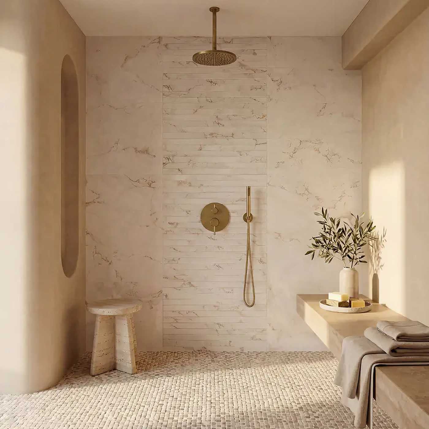 Image of A modern bathroom shower area with warm beige marble wall panels, a round brass rain shower head, matching brass controls, a small stone stool, and a floating shelf with towels and decorative branches.