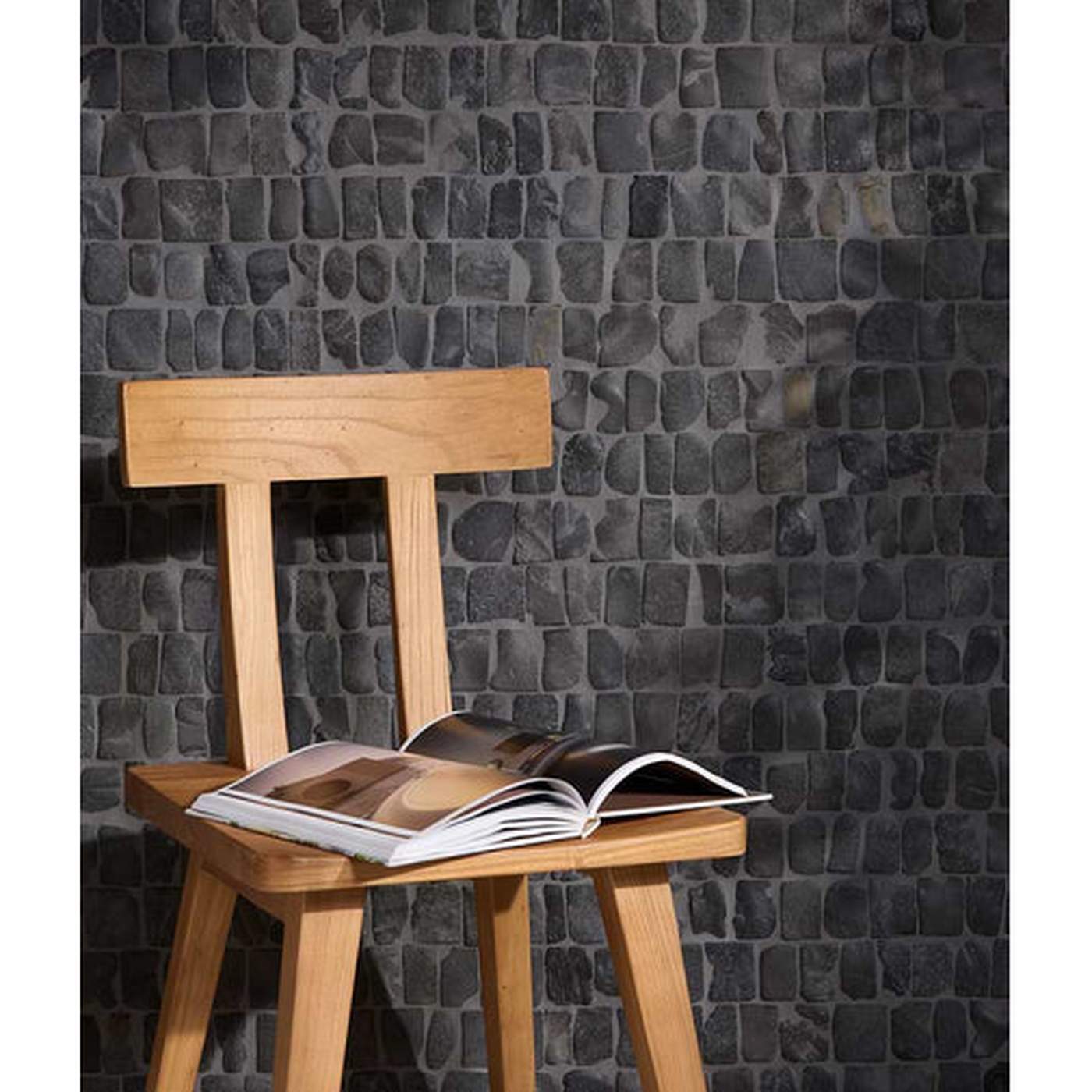 Image of A light natural wood chair with an open magazine resting on its seat, set against a dark gray stone-tile wall.