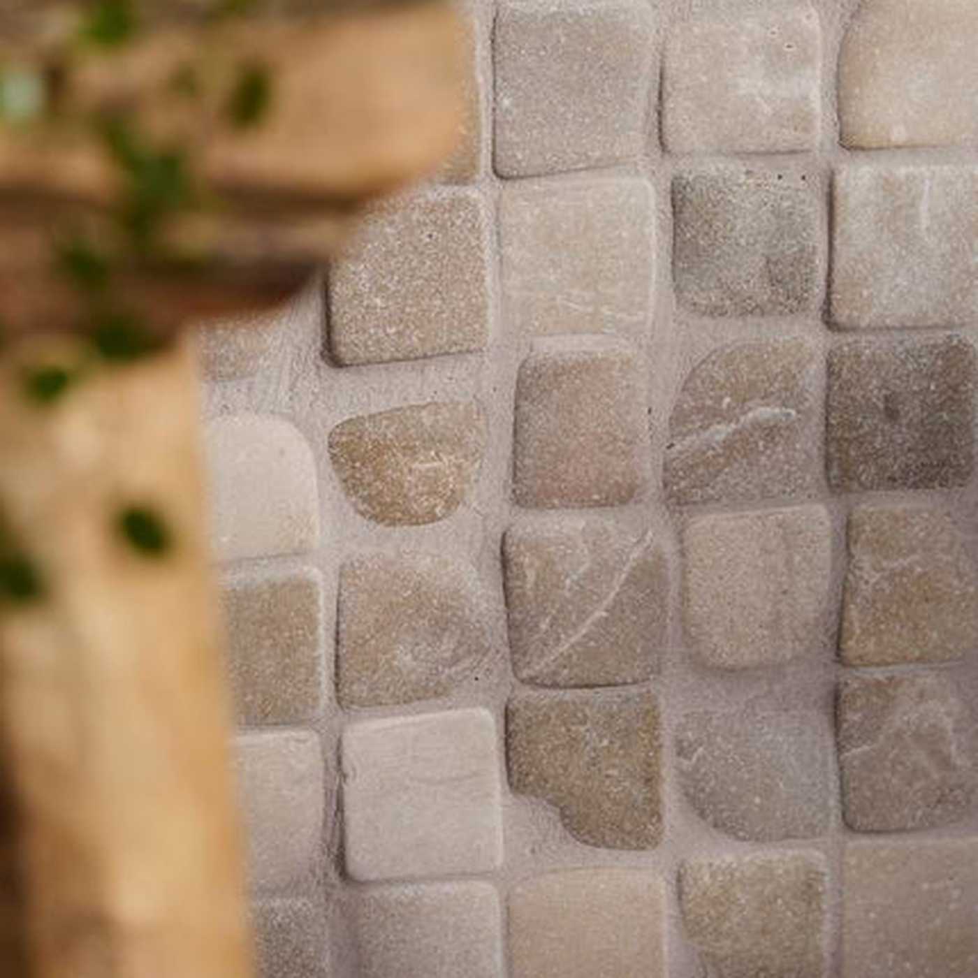 Image of Close-up of a stone mosaic tile wall with irregular square tiles in warm beige, taupe, and light gray tones, with a blurred wooden and green plant element on the left side.