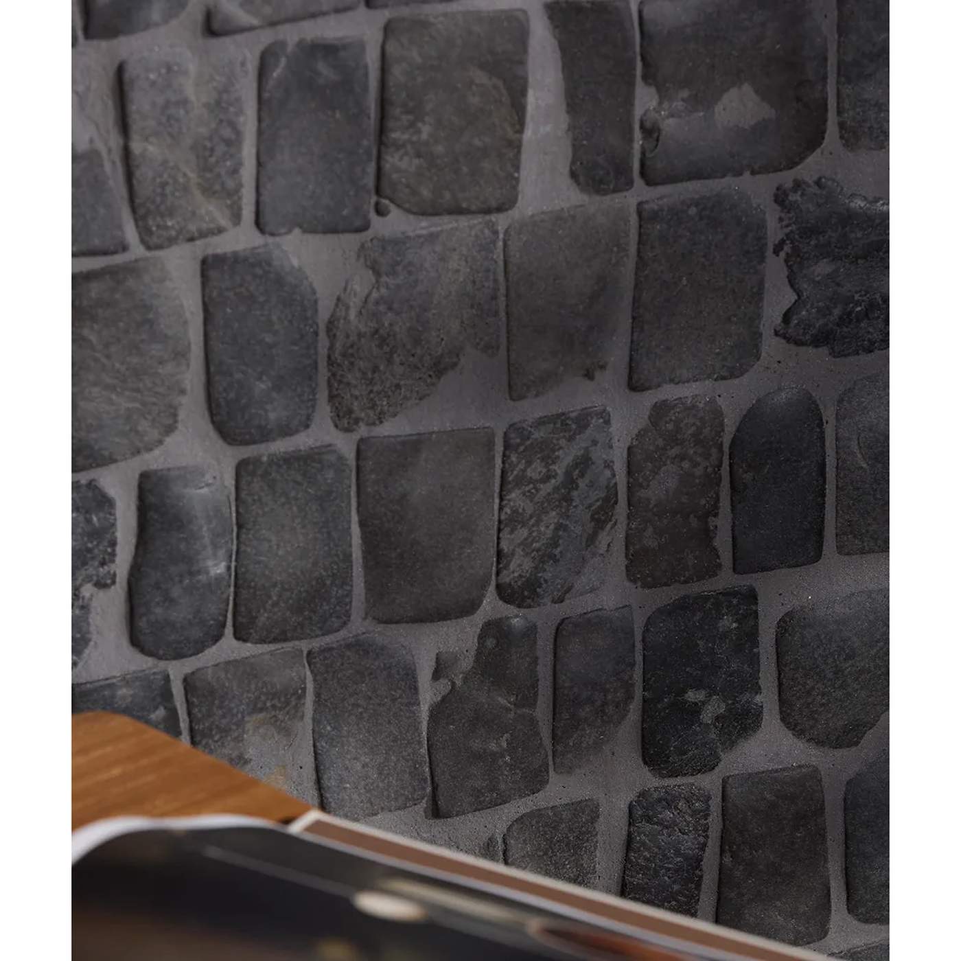 Image of Dark gray stone tile wall with irregular rectangular stones in a mosaic pattern, shown close up with a small portion of a wooden surface visible in the lower left corner.