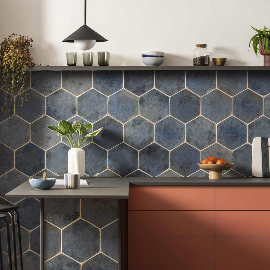 Oken Blue Hexagon tiles 26.7x23.2cm in a kitchen setting with terracotta colour kitchen units, grey worktop featuring apples in a  bowl, vase and breakfast setting. A grey shelf is halfway up the wall with potted plants, glasses, bowls and a jar. Black industrial pendant light hands from the ceiling.
