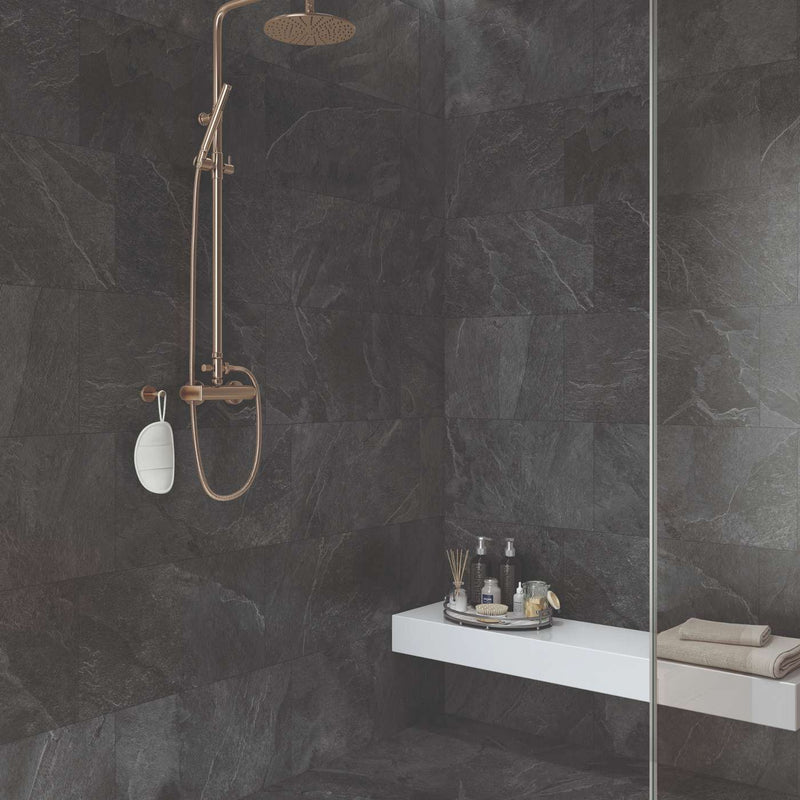 Image of A modern shower cabinetry corner with dark charcoal stone tile walls and floor, featuring a matte bronze shower fixture with a round rain head and handheld wand, a white floating shelf with toiletries, and a clear glass door frame.