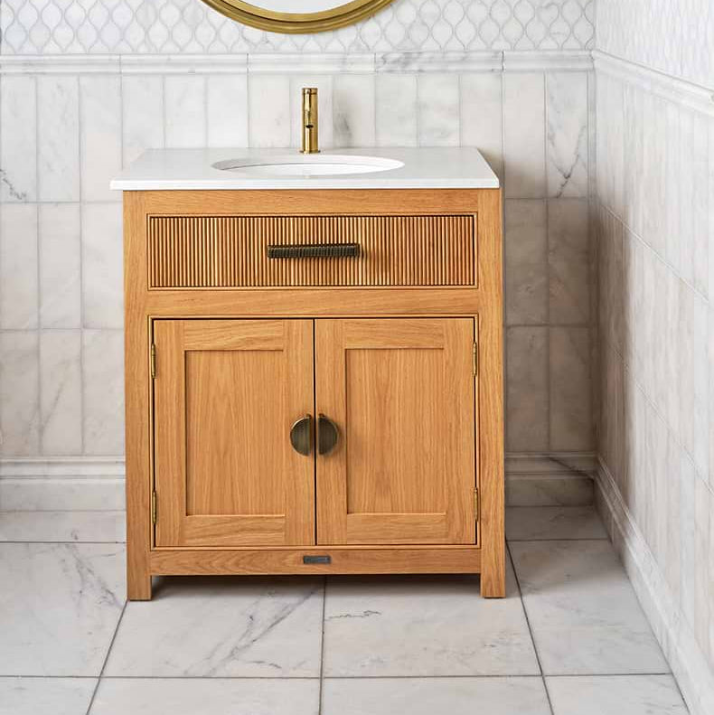 Image of Wooden bathroom vanity with white countertop and round mirror in a tiled bathroom.