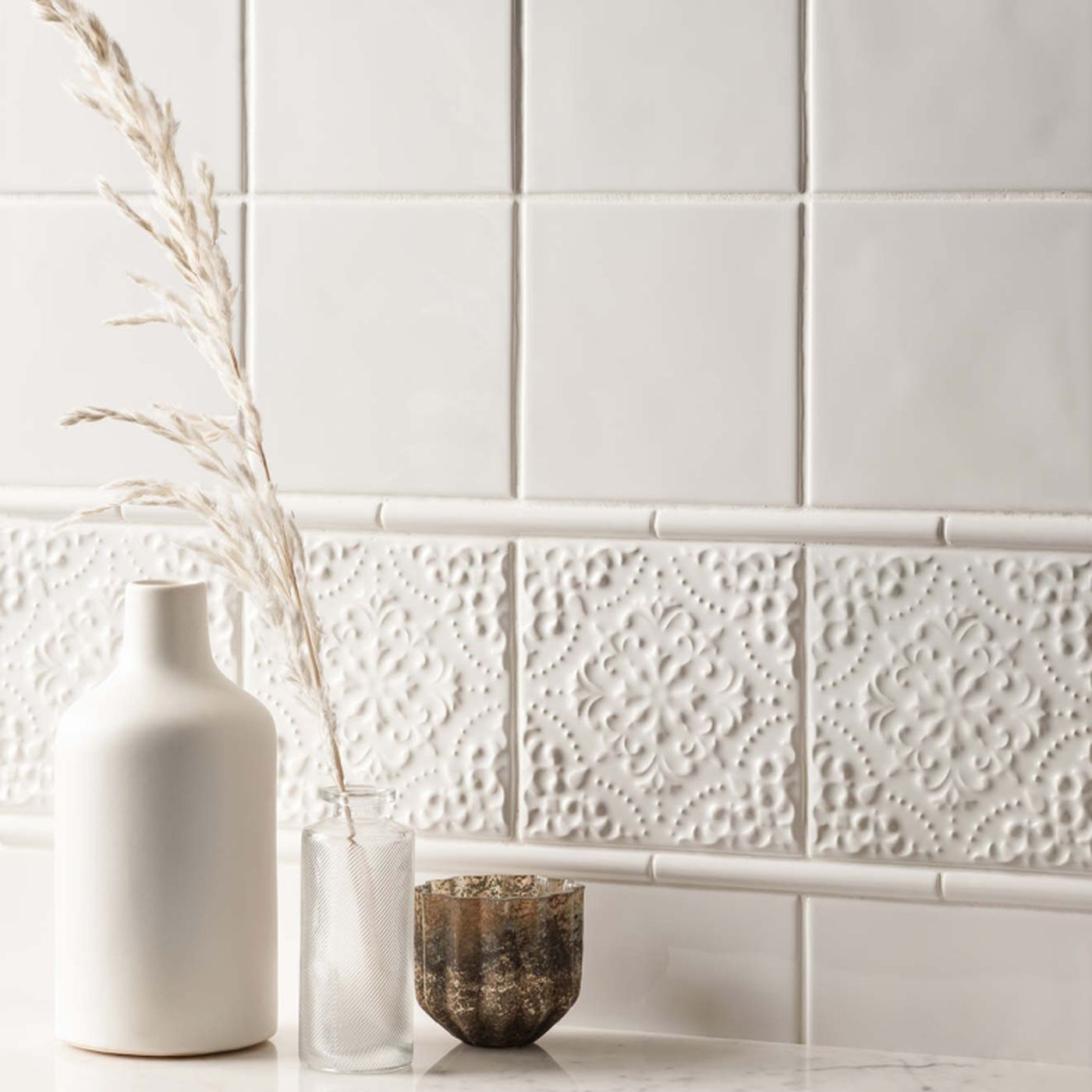Image of White ceramic wall tiles with a decorative embossed border band, styled with a white vase, a clear glass vase with dried grass, and a small textured bowl on a marble surface.
