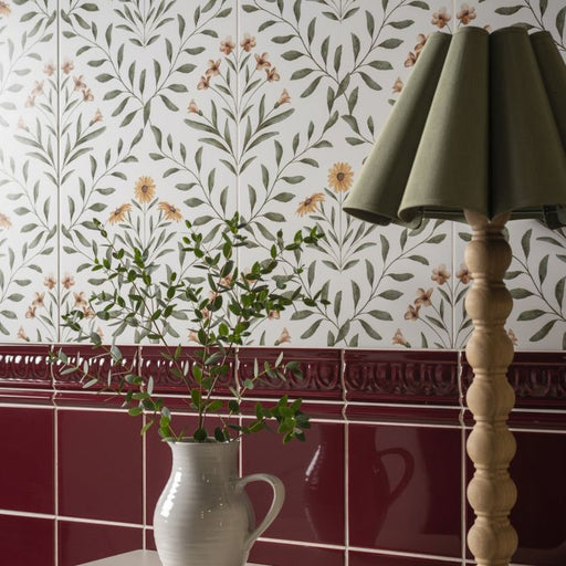 Floral wall tiles with a red border, a white jug of greenery, and a wooden lamp with a green shade.