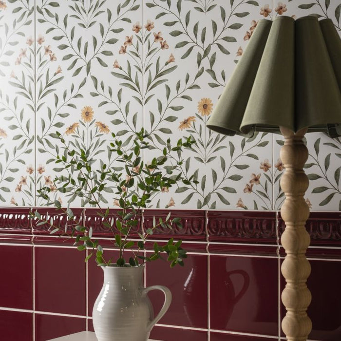 Floral wall tiles with a red border, a white jug of greenery, and a wooden lamp with a green shade.