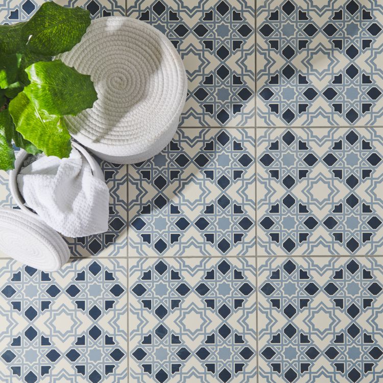 Image of Patterned floor tiles in dark blue and grey with a geometric design, shown alongside white woven baskets and a green plant.”