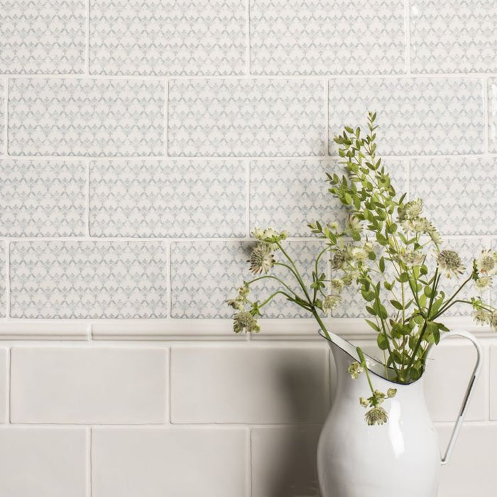 Patterned light blue-and-white wall tiles above glossy white tiles, with a white ceramic jug holding green foliage