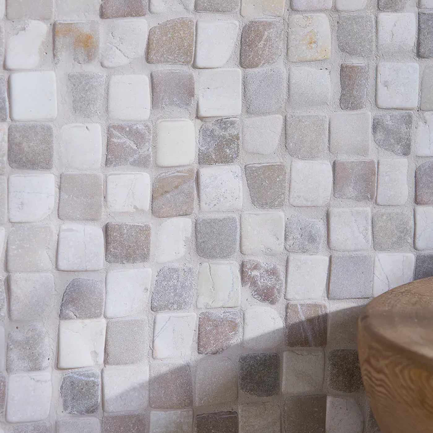 Image of Square mosaic tile wall in neutral stone tones with white, beige, gray, and light brown pieces, shown with a wooden bowl partially visible in the lower right corner.