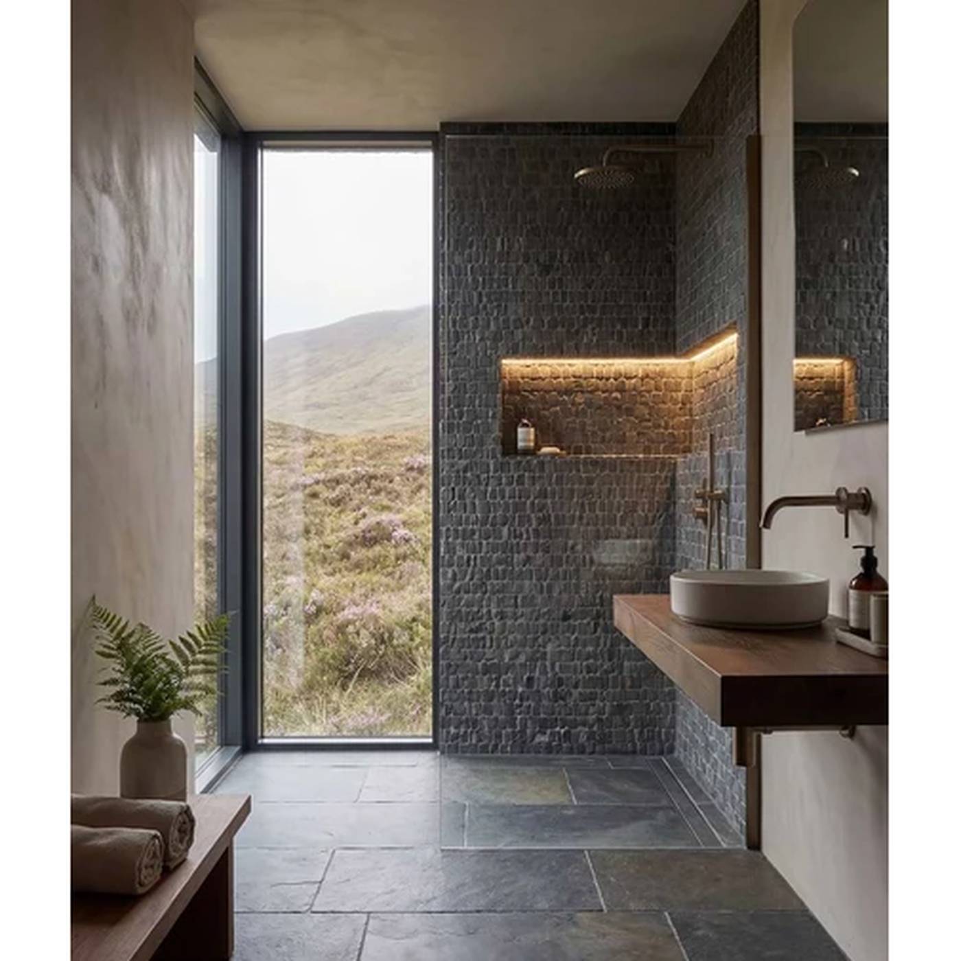 Image of Modern bathroom interior with a stone-tile shower wall, floating wooden vanity, vessel sink, and a large window overlooking a grassy mountain landscape.