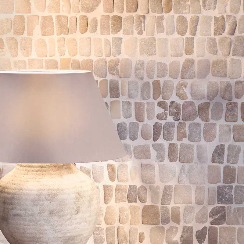 Image of A close-up of a stone mosaic tile wall in neutral beige, cream, and taupe tones, with part of a matching ceramic lamp visible in the lower left corner.