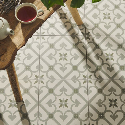 Patterned floor tiles in soft green and grey with a floral trellis design, shown beside a rustic wooden table holding a cup of tea.