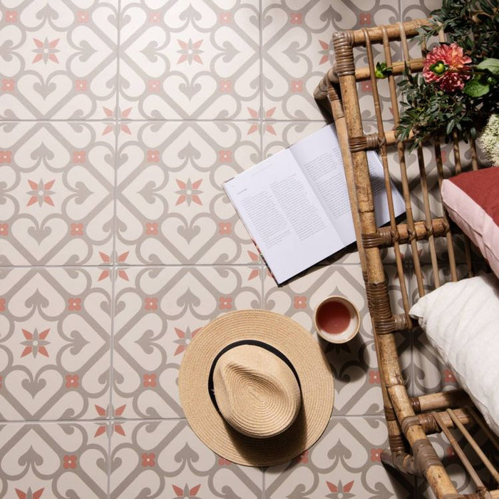Patterned floor tiles in soft grey and rose tones beside a woven bench, with an open book, a cup of tea, and a straw hat placed on the floor