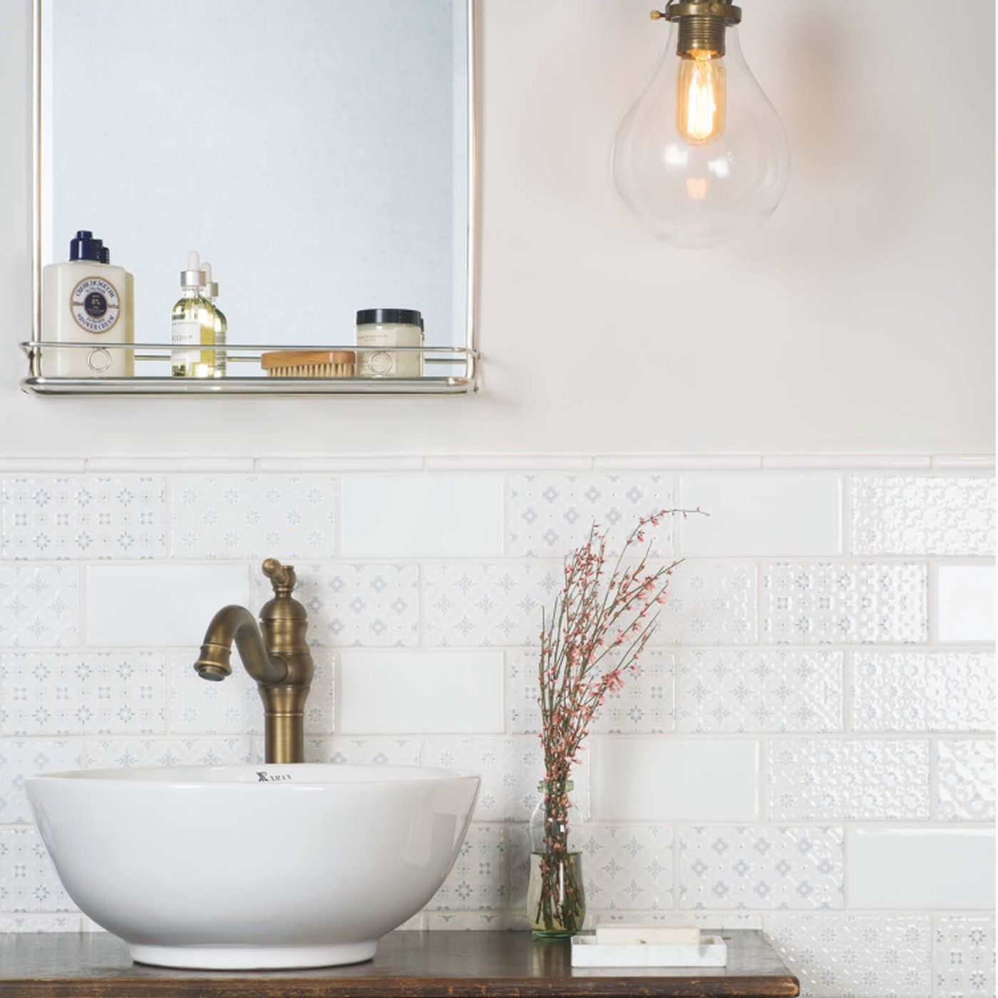 Image of A styled bathroom scene featuring a white ceramic vessel sink with a brass faucet, set against a white patterned tile backsplash. A matching wall mirror with a glass shelf sits above, holding toiletries, and a clear glass pendant light with a warm bulb hangs on the right.