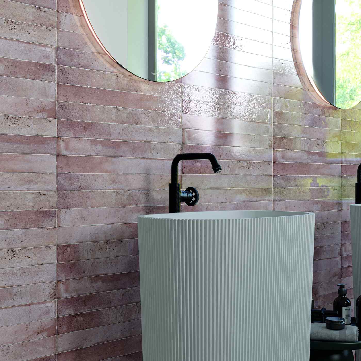 Image of A modern bathroom scene featuring a white ribbed pedestal sink with a matte black faucet, set against a wall of pink textured rectangular tiles. Two round mirrors with copper-toned frames are visible in the background.