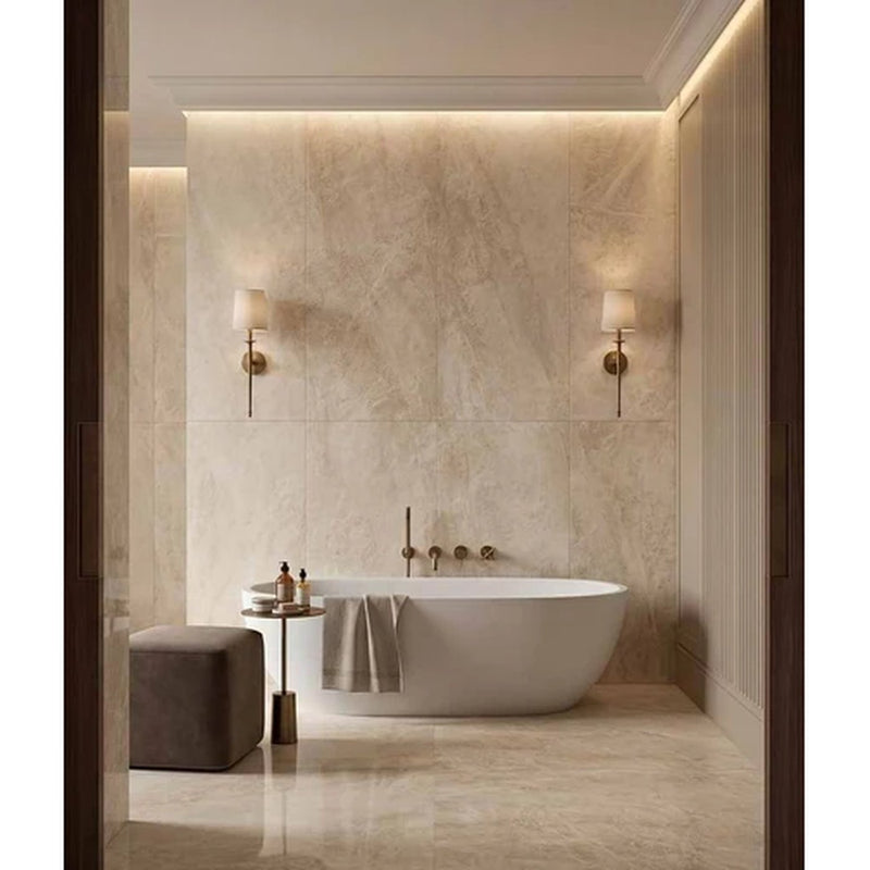 Image of A luxury freestanding oval bathtub in a modern bathroom with beige marble walls and floor, featuring dual brass wall sconces, a small side table, and a gray ottoman.