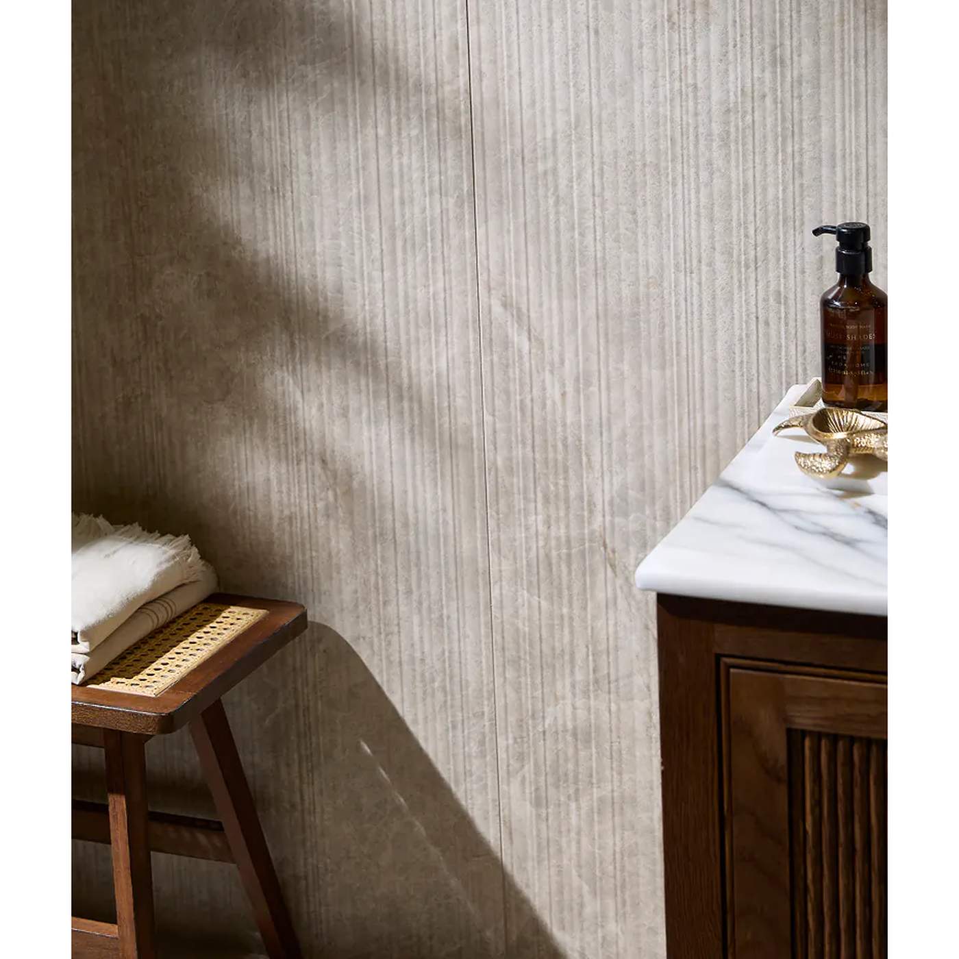 Image of Beige textured wallpaper with vertical ribbed lines, styled in a bathroom setting with a wooden vanity, marble countertop, and a small stool.