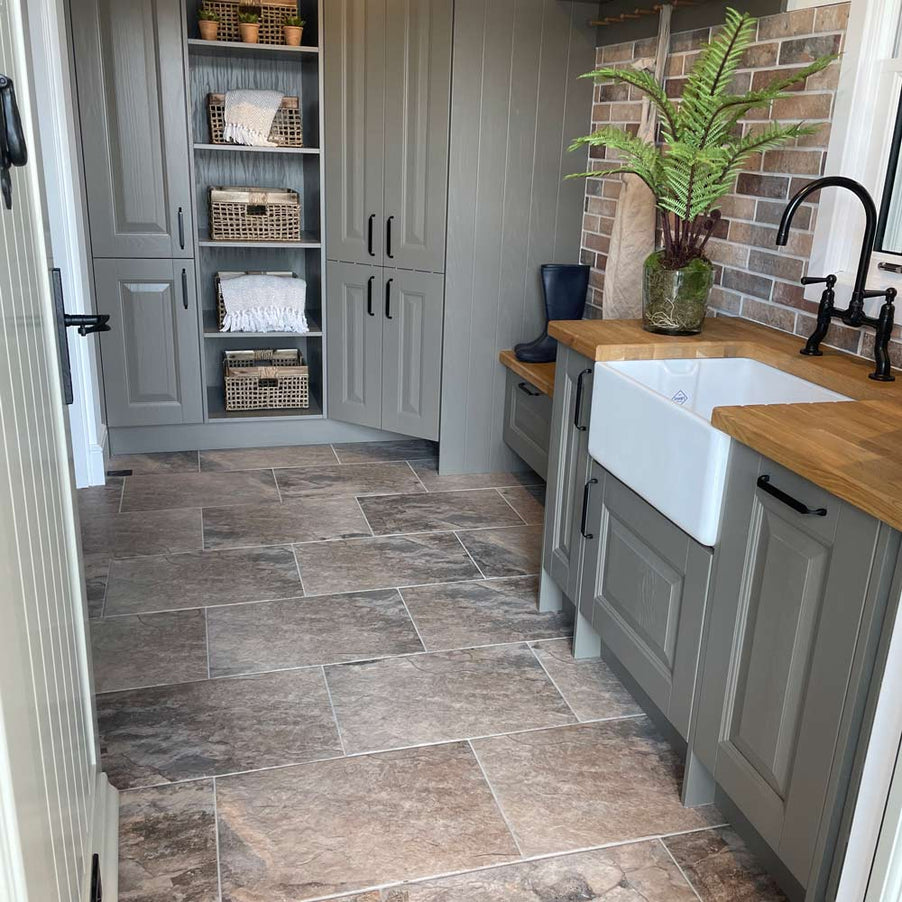 Keystone Stone effect tiles 40x60cm in a kitchen setting. Light grey shaker style kitchen cabinets with wooden worktop, belfast sink and pantry cupboard to the far end. Brick effect tiles on the wall and green plant in a vase near the sink. Cottagecore and country kitchen look
