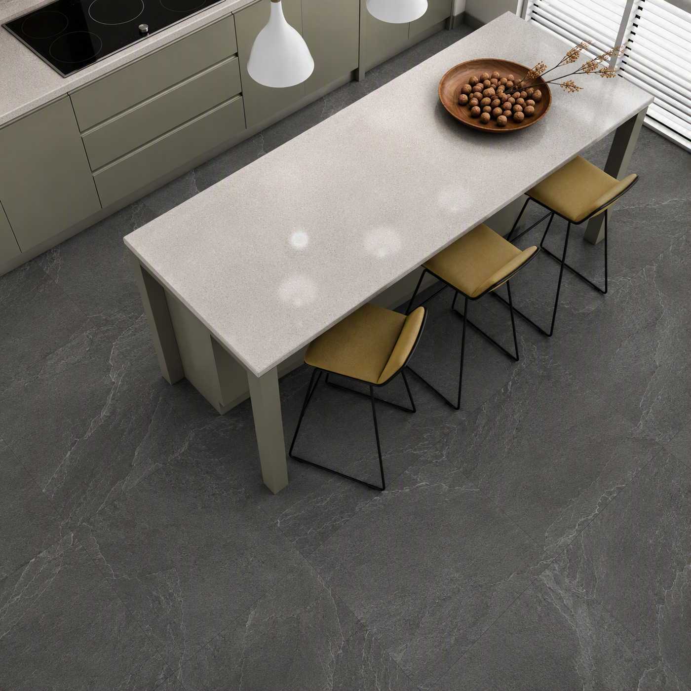Image of A modern kitchen scene with a long rectangular island table in a light stone or concrete finish, surrounded by four yellow upholstered chairs with black metal legs. The room has dark gray stone flooring, light gray cabinetry, pendant lights, and a bowl of nuts on the table.