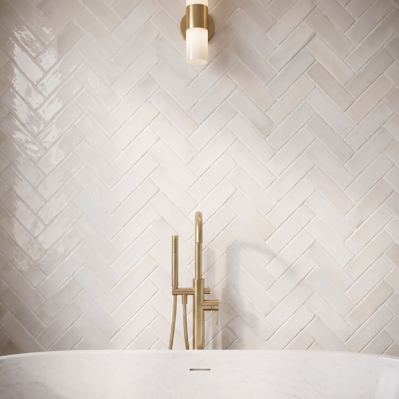 Image of Pearl white herringbone subway tile backsplash behind a white bathtub with a gold faucet, illuminated by a gold wall sconce.