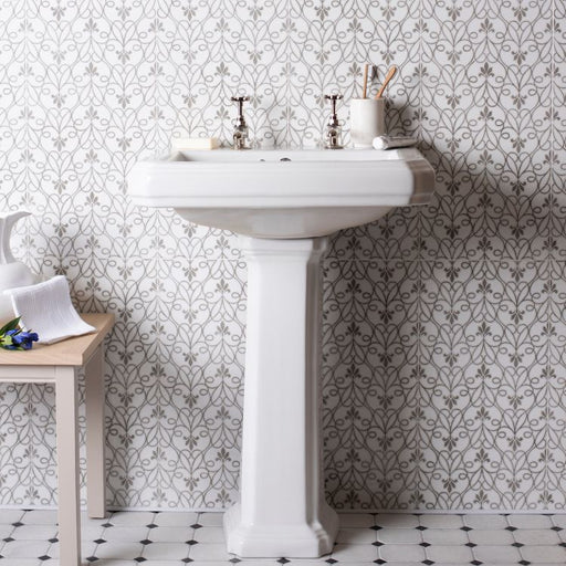 Bathroom with a pedestal sink, patterned grey tile wall, and small wooden side table with decor