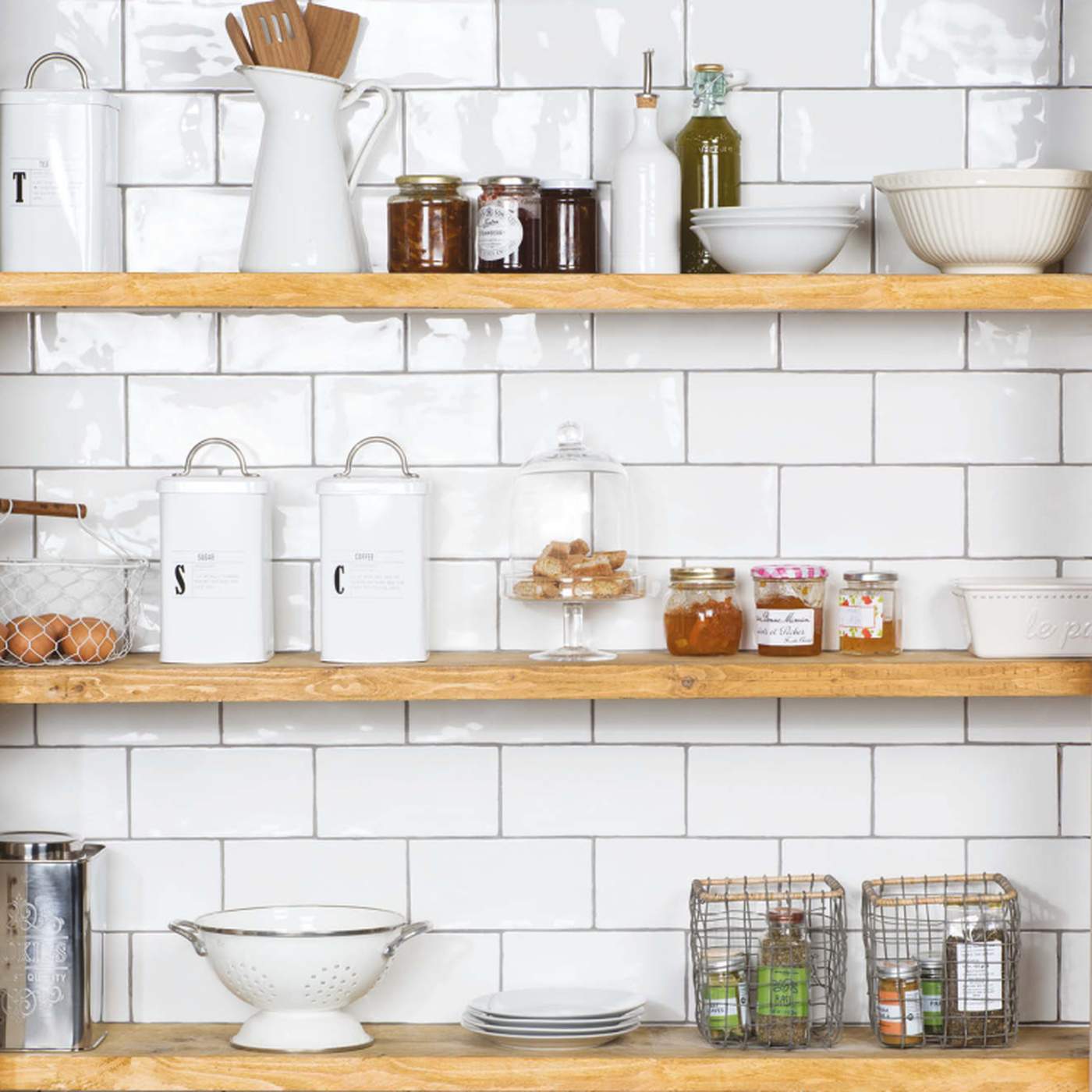 Image of A styled kitchen scene with white subway tile backsplash and three light wood floating shelves holding white ceramic canisters, glass jars, bowls, a pitcher, and wire baskets with small bottles.