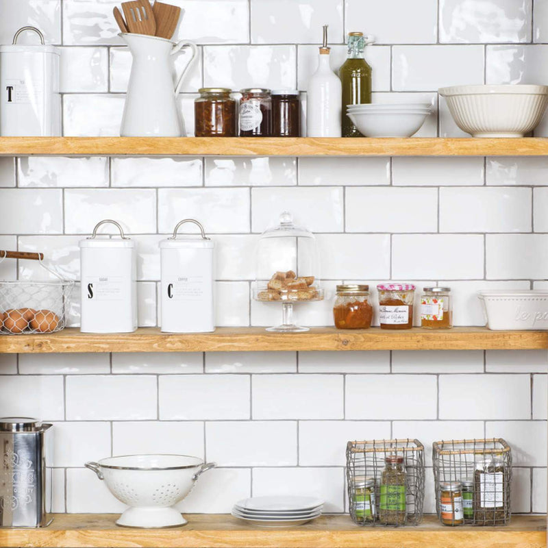 Image of A styled kitchen scene with white subway tile backsplash and three light wood floating shelves holding white ceramic canisters, glass jars, bowls, a pitcher, and wire baskets with small bottles.